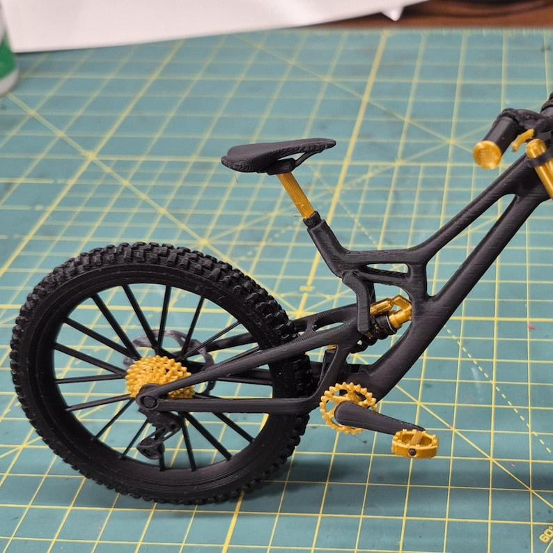 Puede incluir: Una bicicleta de monta&ntilde;a negra en miniatura con detalles dorados. La bicicleta tiene un cuadro, ruedas y neum&aacute;ticos negros, con engranajes, componentes de suspensi&oacute;n y manillar dorados. La tija del sill&iacute;n es dorada y el sill&iacute;n es negro. La bicicleta est&aacute; dise&ntilde;ada para uso todoterreno.