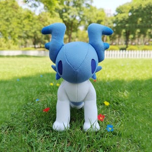Op de afbeelding: Een blauw en wit pluche speelgoed in de vorm van een wezen met grote, gebogen hoorns. Het speelgoed heeft donkerblauwe ogen en staat op groen gras. Het pluche is een zacht, knuffelbaar speelgoed.