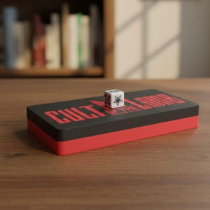 May include: A black and red rectangular box with the text "CULT OF THE LAMB" in red. A white die with a pentagram and star is on top of the box. The box and die are on a wooden surface.