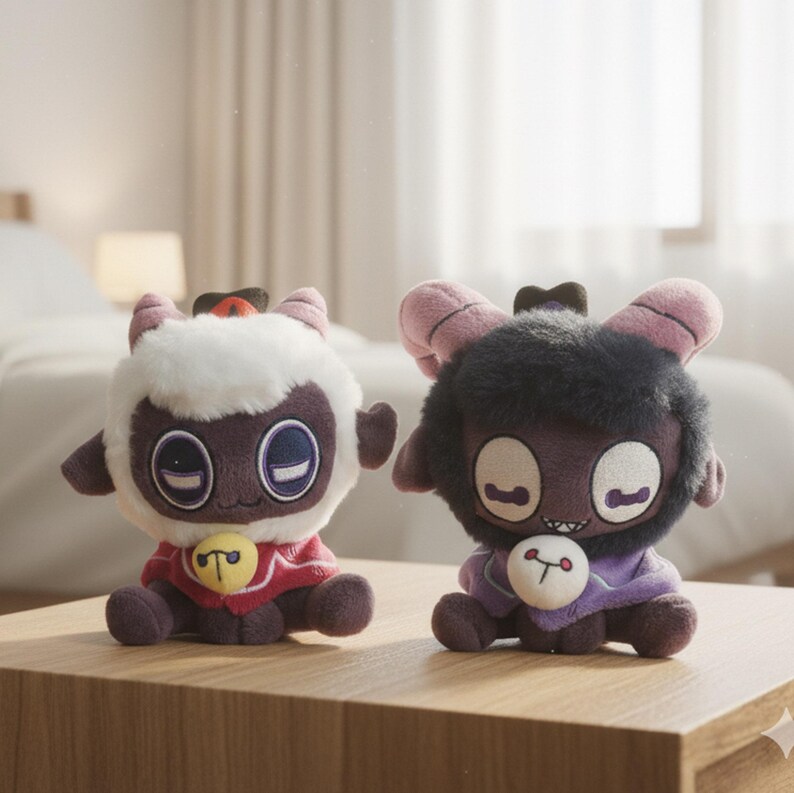 Puede incluir: Dos mu&ntilde;ecos de peluche de cordero est&aacute;n sobre una superficie de madera. Uno es blanco con un cuello rojo y una campana amarilla. El otro es oscuro con un cuello morado y una campana blanca. Ambos tienen ojos grandes y cuernos peque&ntilde;os.