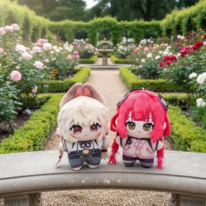 Puede incluir: Dos muñecos de peluche con ojos grandes y trajes detallados. Uno tiene pelo claro y orejas de conejo, el otro tiene pelo rojo. Posan sobre una repisa de piedra en un jardín de rosas con una fuente al fondo.