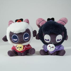 Puede incluir: Dos mu&ntilde;ecos de peluche de cordero con ojos grandes, cuernos y peque&ntilde;as capas. Uno tiene pelaje blanco y una capa roja, el otro tiene pelaje oscuro y una capa morada. Ambos tienen un bot&oacute;n con un s&iacute;mbolo.