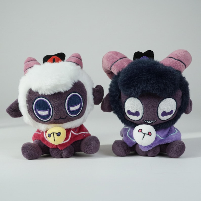 Puede incluir: Dos mu&ntilde;ecos de peluche de cordero con ojos grandes, cuernos y peque&ntilde;as capas. Uno tiene pelaje blanco y una capa roja, el otro tiene pelaje oscuro y una capa morada. Ambos tienen un bot&oacute;n con un s&iacute;mbolo.