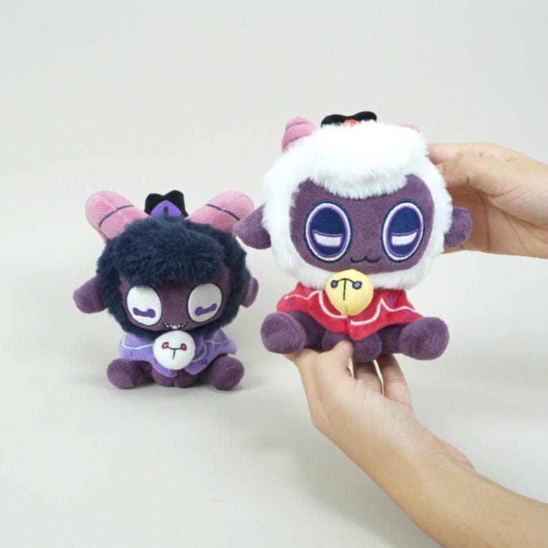 Puede incluir: Dos peluches de un cordero de dibujos animados con cuerpos morados, ojos grandes y cuernos peque&ntilde;os. Un cordero tiene una cabeza blanca y esponjosa y un cuello rojo con una campana dorada. El otro cordero tiene una cabeza azul oscuro y un atuendo morado.