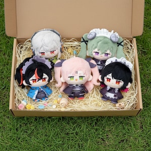 Op de afbeelding: Vijf pluche poppen in anime-stijl zijn gerangschikt in een kartonnen doos gevuld met stro. De poppen hebben verschillende haarkleuren, waaronder wit, groen, zwart en roze. Elke pop heeft een unieke outfit en gezichtsuitdrukking, met grote ogen en kleine monden.