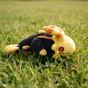 May include: A plush toy featuring a black body with yellow accents, including long ears with orange stripes, a yellow head, and a small tail. The creature has a round pink eye and a stitched mouth, crafted from soft fabric in a reclining position.