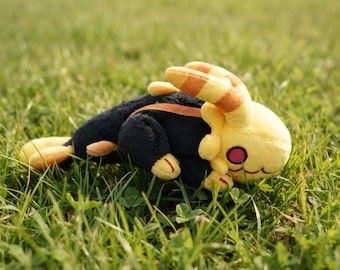 May include: A plush toy featuring a black body with yellow accents, including long ears with orange stripes, a yellow head, and a small tail. The creature has a round pink eye and a stitched mouth, crafted from soft fabric in a reclining position.