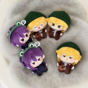 May include: A collection of plush dolls. Some dolls have yellow hair and wear green hats and brown outfits, holding small guitars. Other dolls have purple hair and frog-shaped headbands. The dolls are on a soft, gray surface.