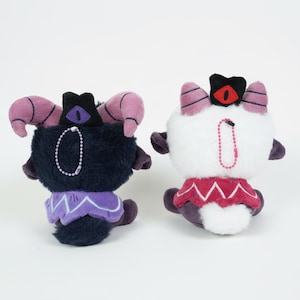 Puede incluir: Dos peluches, uno negro y otro blanco, con dise&ntilde;os similares. Cada uno tiene cuernos, una corona y un cuello decorativo. El juguete negro tiene un cuello morado, mientras que el juguete blanco tiene un cuello rojo. Ambos tienen una cadena.