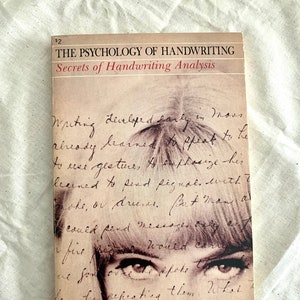 May include: A paperback book titled The Psychology of Handwriting: Secrets of Handwriting Analysis by Nadya Olyanova. The cover features a sepia-toned close-up of a person's eyes overlaid with cursive handwriting text against a light background.