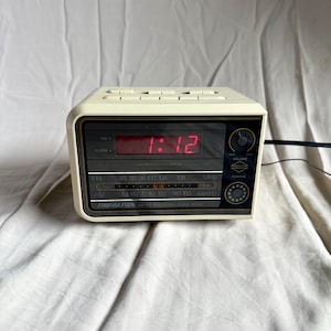 May include: A vintage cream-colored Soundesign digital clock radio. The clock displays the time 1:12 in red digits. It has a black face with tuning and volume controls, and an AM/FM radio dial.