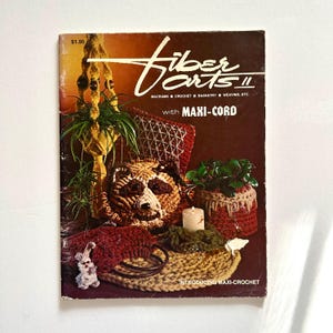 May include: A vintage instructional book titled Fiber Arts II with Maxi-Cord, featuring a dark brown cover with images of various macrame and crochet projects, including a raccoon face, a hanging plant holder, and a small white candle.