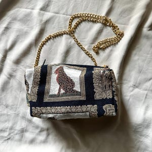 May include: A small, rectangular tapestry bag with a gold chain strap. The bag features a detailed image of a seated dog in a framed design. The background is navy blue with a repeating pattern. The chain is a bright gold color.