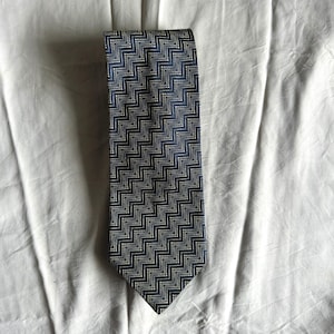 Puede incluir: Una corbata azul marino con un patrón de zigzag repetido en blanco y azul claro. La corbata está hecha de una tela suave y se muestra sobre un fondo blanco. Un accesorio clásico para vestimenta formal o de negocios.