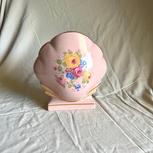 May include: A pink ceramic shell-shaped vase with a floral bouquet design. The vase has a scalloped edge and a rectangular base. The floral bouquet includes yellow, pink, and blue flowers with green leaves. Gold trim accents the edges.