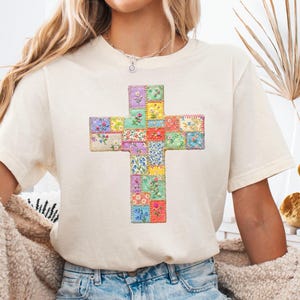 Könnte beinhalten: Cremefarbenes T-Shirt mit einem farbenfrohen Kreuzdesign. Das Kreuz besteht aus kleinen, quadratischen Patches mit Blumenmustern in verschiedenen Farben, darunter Rosa, Blau, Gelb und Grün. Das Shirt wird mit einer blauen Jeans kombiniert.