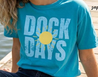 Puede incluir: Una camiseta de manga corta color azul zafiro con el texto DOCK DAYS en una fuente azul claro desgastada. Un gráfico de sol amarillo sólido está posicionado en el centro del texto. La camiseta tiene cuello redondo y corte estándar.