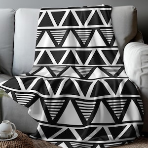 May include: A black and white geometric patterned throw blanket draped over a gray sofa. The blanket features a repeating design of triangles and horizontal lines. The throw is made of a soft material.