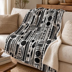 May include: A plush throw blanket featuring a black and white pattern of vertical stripes composed of various geometric shapes, circles, and abstract line art. The blanket has a soft, textured white fleece lining visible on the folded corner.