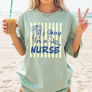 Op de afbeelding: Saliegroen T-shirt met de tekst "It's Okay I'm a Nurse" in blauwe letters en een stethoscoopafbeelding. De tekst staat tegen een achtergrond van gele verticale strepen. Het shirt heeft een relaxte pasvorm.
