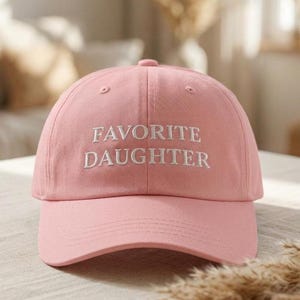 May include: A pale pink baseball cap featuring the embroidered words "FAVORITE DAUGHTER" in white. The cap has a curved peak and a rounded crown. The background is blurred, suggesting an indoor setting.