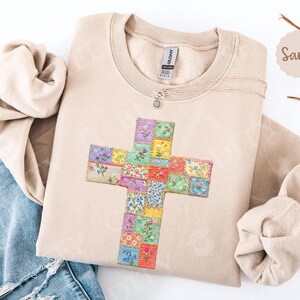 Puede incluir: Sudadera color arena con un dise&ntilde;o de cruz de patchwork floral. La cruz est&aacute; hecha de peque&ntilde;os cuadrados coloridos con varios patrones de flores. Se ve un collar plateado con un peque&ntilde;o colgante. La sudadera est&aacute; doblada junto a unos vaqueros azules.