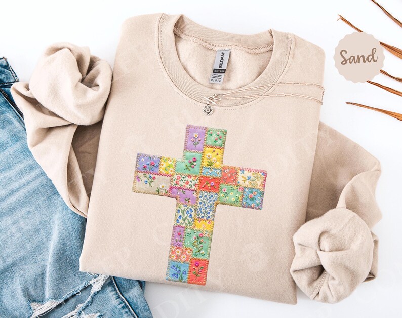 Puede incluir: Sudadera color arena con un dise&ntilde;o de cruz de patchwork floral. La cruz est&aacute; hecha de peque&ntilde;os cuadrados coloridos con varios patrones de flores. Se ve un collar plateado con un peque&ntilde;o colgante. La sudadera est&aacute; doblada junto a unos vaqueros azules.