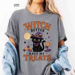 May include: A grey t-shirt featuring a graphic of a black cat wearing a purple witch hat and bow tie. The text reads "WITCH BETTER HAVE MY TREATS" in orange letters. The design includes a yellow moon, stars, and colorful Halloween candy.