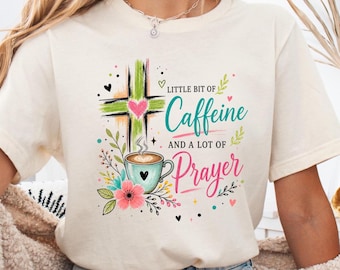 Puede incluir: Una camiseta color crema con un diseño gráfico que incluye una cruz verde, un corazón rosa, una taza de café azul claro y detalles florales. El texto dice: Little bit of Caffeine AND A LOT OF Prayer, con Caffeine y Prayer en cursiva.