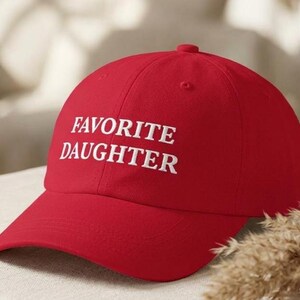 May include: A red baseball cap with the text "FAVORITE DAUGHTER" embroidered in white. The cap features a curved peak and a rounded crown. The hat is made of a soft material and is suitable for casual wear.