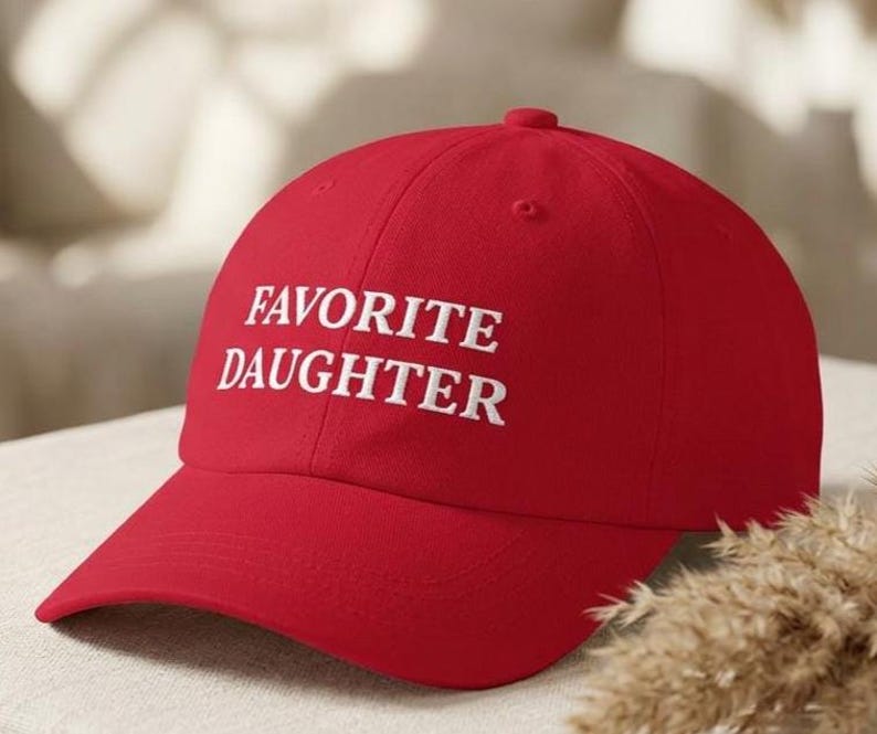 May include: A red baseball cap with the text "FAVORITE DAUGHTER" embroidered in white. The cap features a curved peak and a rounded crown. The hat is made of a soft material and is suitable for casual wear.
