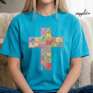Puede incluir: Una camiseta turquesa con un dise&ntilde;o de cruz colorida. La cruz est&aacute; compuesta por cuadrados de tela estampada floral en tonos rojos, amarillos, verdes y azules. La palabra "sapphire" est&aacute; escrita en cursiva en la parte superior derecha.