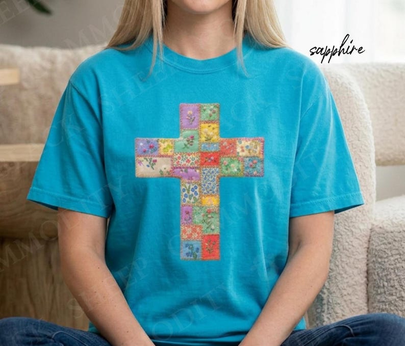 Puede incluir: Una camiseta turquesa con un dise&ntilde;o de cruz colorida. La cruz est&aacute; compuesta por cuadrados de tela estampada floral en tonos rojos, amarillos, verdes y azules. La palabra "sapphire" est&aacute; escrita en cursiva en la parte superior derecha.