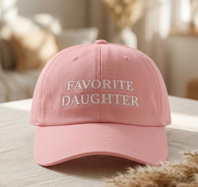 May include: A pale pink baseball cap featuring the embroidered words "FAVORITE DAUGHTER" in white. The cap has a curved peak and a rounded crown. The background is blurred, suggesting an indoor setting.