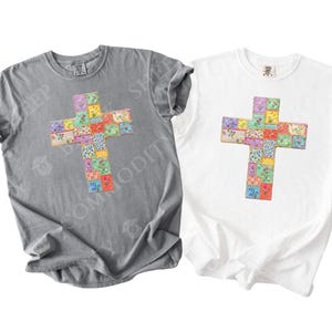 Puede incluir: Dos camisetas, una gris y otra blanca, cada una con un dise&ntilde;o de cruz colorida. La cruz est&aacute; compuesta por varios parches cuadrados con dise&ntilde;os florales y estampados. La palabra "grey" est&aacute; escrita en cursiva a la izquierda y "white" a la derecha.