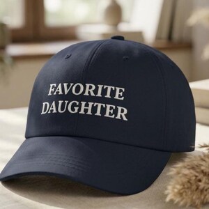May include: A navy blue baseball cap featuring the text "FAVORITE DAUGHTER" in white embroidery. The cap has a curved peak and a structured crown, ideal for everyday use.