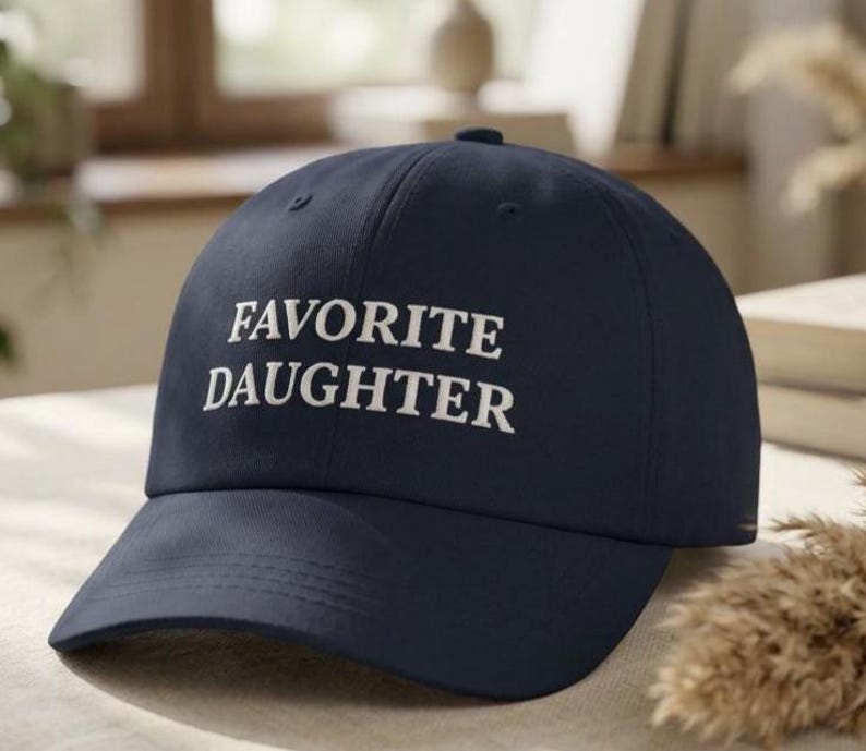 May include: A navy blue baseball cap featuring the text "FAVORITE DAUGHTER" in white embroidery. The cap has a curved peak and a structured crown, ideal for everyday use.