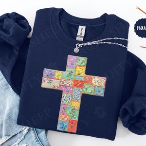 Puede incluir: Sudadera azul marino con un dise&ntilde;o de cruz colorida. La cruz est&aacute; hecha de cuadrados de patchwork con estampados florales. Un collar plateado con un peque&ntilde;o colgante est&aacute; sobre la sudadera.
