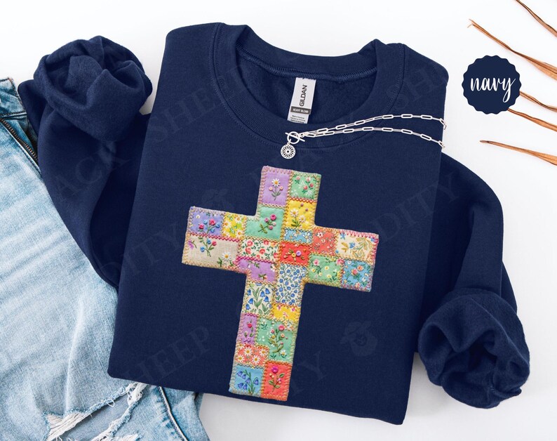 Puede incluir: Sudadera azul marino con un dise&ntilde;o de cruz colorida. La cruz est&aacute; hecha de cuadrados de patchwork con estampados florales. Un collar plateado con un peque&ntilde;o colgante est&aacute; sobre la sudadera.