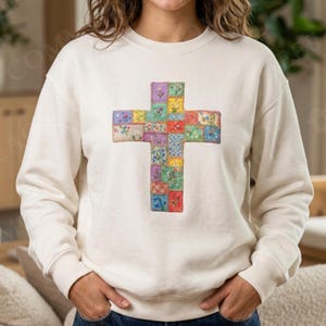 Puede incluir: Sudadera color crema con un dise&ntilde;o de cruz de patchwork colorido. La cruz est&aacute; compuesta por varios cuadrados de tela en tonos rojos, azules, morados y amarillos. La sudadera tiene cuello redondo y mangas largas.