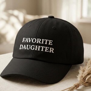 May include: Black baseball cap with the embroidered text "FAVORITE DAUGHTER" in white. The cap has a curved peak and a button on top. The hat is displayed on a neutral surface.