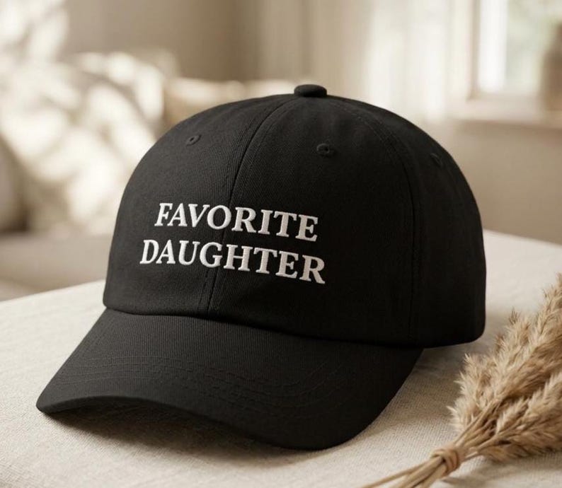 May include: Black baseball cap with the embroidered text "FAVORITE DAUGHTER" in white. The cap has a curved peak and a button on top. The hat is displayed on a neutral surface.