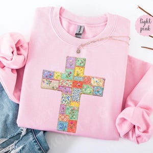 Puede incluir: Sudadera rosa claro con un dise&ntilde;o de cruz. La cruz est&aacute; hecha de coloridos cuadrados de patchwork floral. Un delicado collar con un peque&ntilde;o colgante descansa sobre la sudadera. La palabra "light pink" est&aacute; escrita en un c&iacute;rculo rosa.