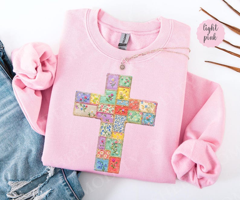 Puede incluir: Sudadera rosa claro con un dise&ntilde;o de cruz. La cruz est&aacute; hecha de coloridos cuadrados de patchwork floral. Un delicado collar con un peque&ntilde;o colgante descansa sobre la sudadera. La palabra "light pink" est&aacute; escrita en un c&iacute;rculo rosa.