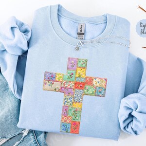 Puede incluir: Sudadera azul claro con un dise&ntilde;o de cruz de patchwork floral. La cruz est&aacute; hecha de cuadrados de tela con flores bordadas en m&uacute;ltiples colores. Un collar plateado con un peque&ntilde;o colgante est&aacute; sobre la sudadera.