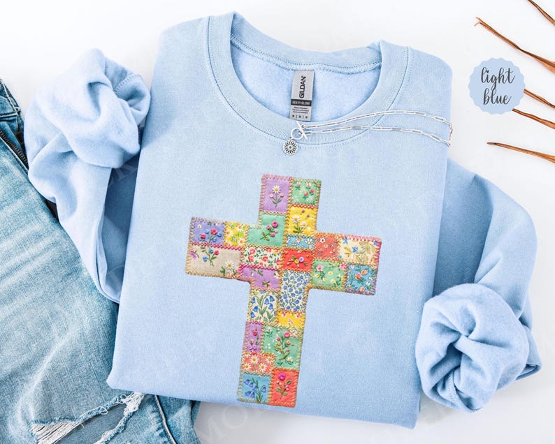 Puede incluir: Sudadera azul claro con un dise&ntilde;o de cruz de patchwork floral. La cruz est&aacute; hecha de cuadrados de tela con flores bordadas en m&uacute;ltiples colores. Un collar plateado con un peque&ntilde;o colgante est&aacute; sobre la sudadera.
