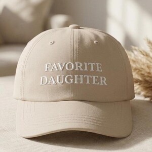 May include: A beige baseball cap with the embroidered text "FAVORITE DAUGHTER" in white. The cap has a curved brim and a button on top. The hat is a neutral colour and suitable for everyday wear.