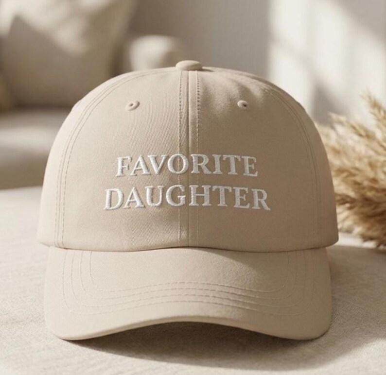 May include: A beige baseball cap with the embroidered text "FAVORITE DAUGHTER" in white. The cap has a curved brim and a button on top. The hat is a neutral colour and suitable for everyday wear.