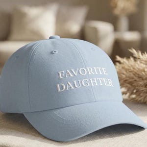 May include: A pale blue baseball cap featuring the embroidered words "FAVORITE DAUGHTER" in white. The cap has a curved peak and a rounded crown. The hat is made of a soft fabric.