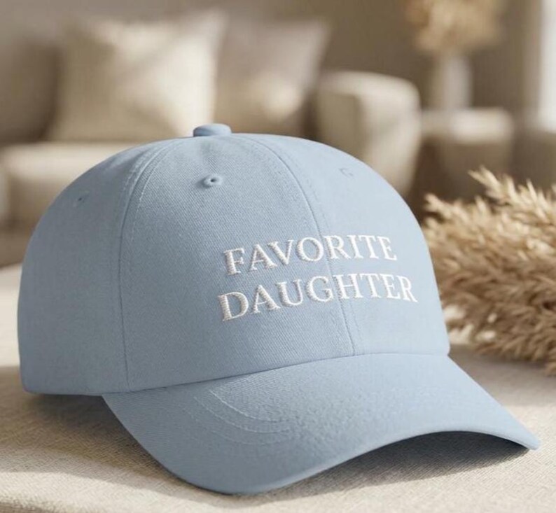 May include: A pale blue baseball cap featuring the embroidered words "FAVORITE DAUGHTER" in white. The cap has a curved peak and a rounded crown. The hat is made of a soft fabric.